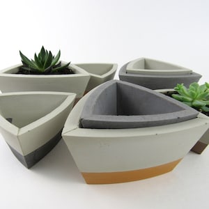 May include: Set of five concrete planters in a geometric triangular shape. The planters are in shades of gray and white with a light orange accent. Two of the planters have small succulents growing in them.