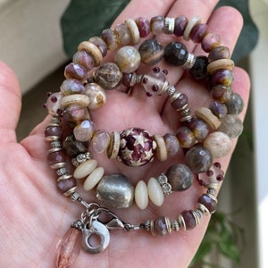 May include: A multi-strand bracelet featuring a variety of beads in shades of purple, tan, and silver. The beads include faceted, round, and cylindrical shapes, with a central bead adorned with a star pattern. The bracelet is secured with a silver clasp.