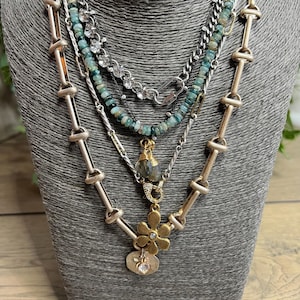 May include: A layered necklace set displayed on a gray bust. The necklaces feature various chains, including silver and gold-toned options. One necklace has small, light blue beads, while another has a flower pendant with a heart charm.