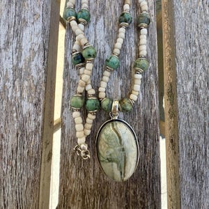 May include: A handcrafted necklace featuring a large oval pendant with green and cream-colored stone. The necklace has a beaded design with cream-colored and green beads, and silver-toned accents. The necklace is displayed on a wooden surface.