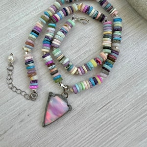 May include: A colorful necklace featuring a pendant and a beaded design. The pendant is a pink, triangular shape with a silver-colored metal frame. The necklace is made of small, round, multi-colored beads in pastel shades.