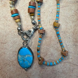 May include: A handcrafted necklace featuring a turquoise pendant and a mix of beads in shades of brown, blue, and silver. The necklace has a unique design with a variety of textures and shapes, creating a bohemian style.