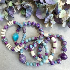 May include: A collection of four stretch bracelets featuring a variety of beads in shades of purple, light blue, turquoise, and cream. The beads include round, cylindrical, and disc shapes with marbled, solid, and striped patterns on elastic cord.