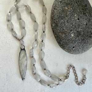May include: A light gray necklace with a long, oval pendant and faceted beads. The pendant is framed in silver and hangs from a beaded strand. A silver chain extends from the necklace. A gray stone is in the background.