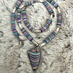 May include: A colorful necklace with a pendant. The necklace features a series of small, round, striped beads in shades of purple, green, blue, and pink, connected by silver-colored metal tubes. The pendant is a triangular shape with a similar striped pattern.