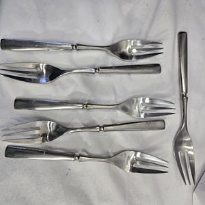 May include: Six silver forks with a silver fork on the right side of the image. The forks have a simple design with a rounded handle and three prongs.