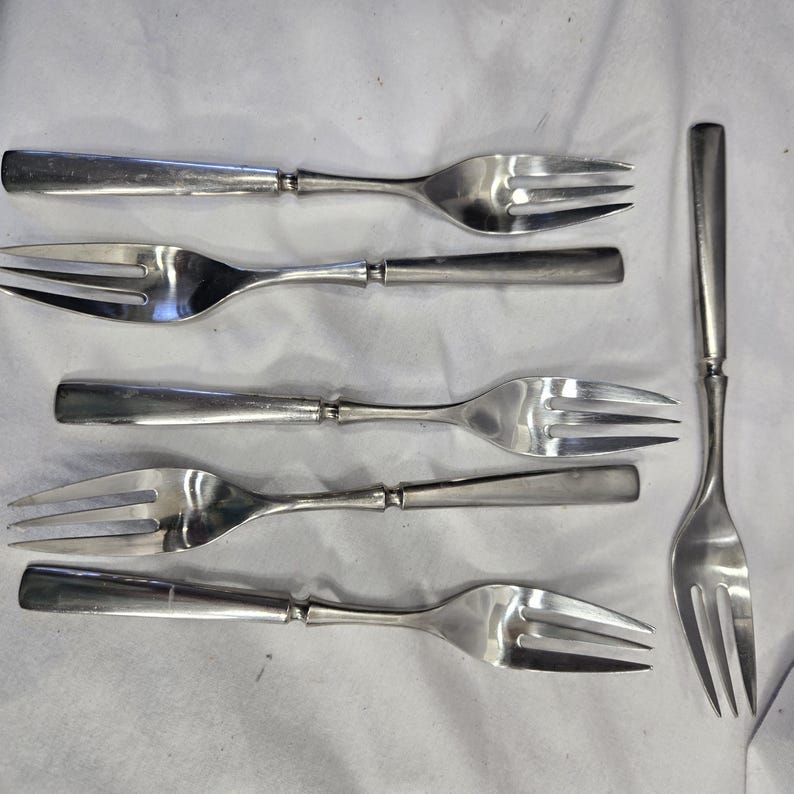 May include: Six silver forks with a silver fork on the right side of the image. The forks have a simple design with a rounded handle and three prongs.