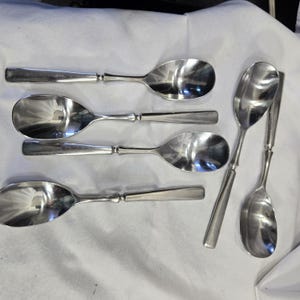 May include: A set of six silver spoons with a rounded bowl shape. The spoons are arranged in a fan shape on a white surface.