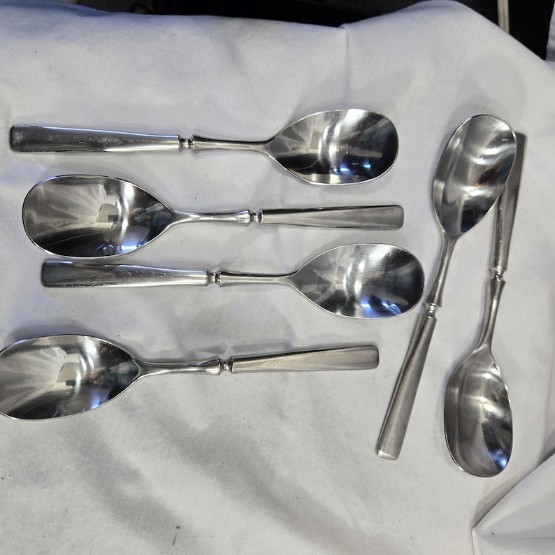 May include: A set of six silver spoons with a rounded bowl shape. The spoons are arranged in a fan shape on a white surface.