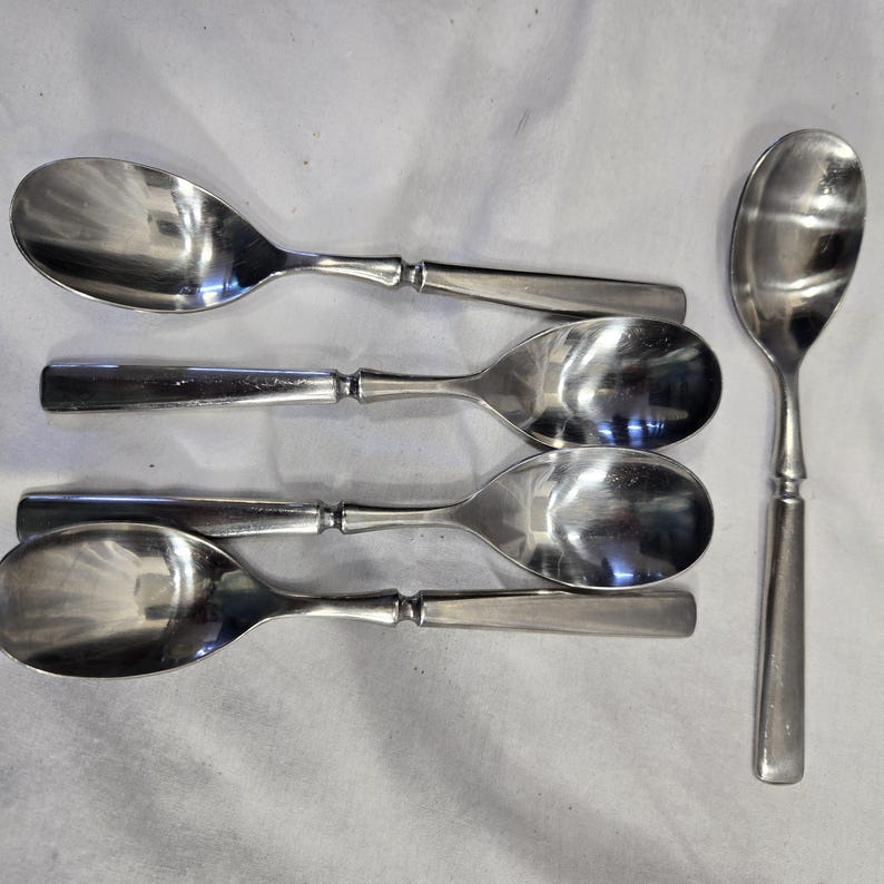 May include: Five silver spoons with rounded bowls and simple handles. The spoons are arranged in a row, with the handles pointing to the right.