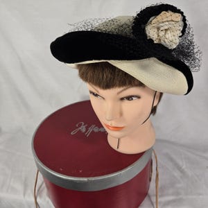 May include: A vintage hat with a black velvet brim and a cream-colored crown. The hat features a black netting veil and a cream-colored fabric flower. The hat is displayed on a mannequin head, atop a red hat box.