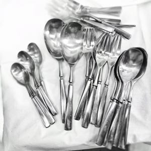 May include: A collection of silver spoons and forks arranged on a white surface. The utensils are all polished and shiny, reflecting the light.