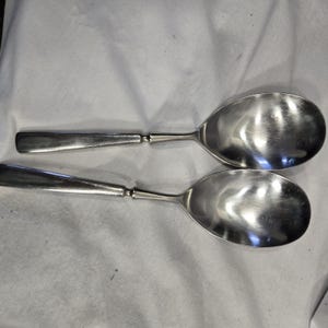 May include: Two silver serving spoons with a shiny finish. The spoons are laid out on a white fabric surface.