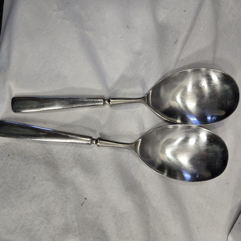 May include: Two silver serving spoons with a shiny finish. The spoons are laid out on a white fabric surface.