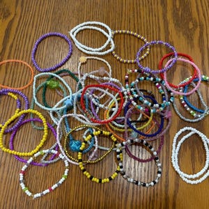 May include: A collection of beaded bracelets in various colors, including purple, yellow, white, red, and green. The bracelets are made of small beads and are arranged on a wooden surface. The bracelets are a mix of solid colors and patterns.