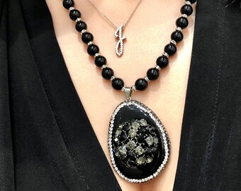 May include: A two-strand necklace featuring a large black pendant with a crystal-studded border and center. The necklace has black beads with silver accents. A smaller necklace with a crystal-studded letter "J" is also visible.