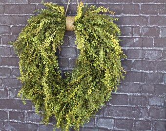May include: A green leafy wreath hanging on a brick wall. The wreath is made of artificial greenery and is tied with twine.