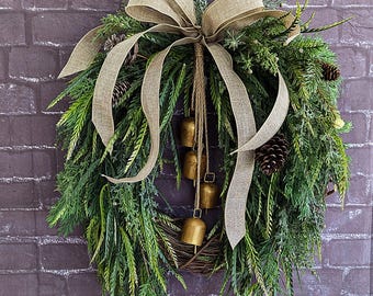 May include: A festive Christmas wreath with green foliage, pinecones, and gold bells. A large burlap bow sits at the top, with long flowing ribbons. The wreath is hanging against a brick wall, creating a rustic holiday decoration.