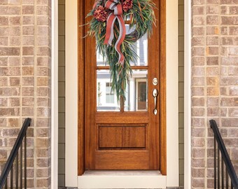 Può includere: Una ghirlanda decorativa con fiori di ortensia rosso scuro, lungo fogliame verde e un fiocco a righe grigie e rosse, appesa a una porta d'ingresso in legno con pannelli in vetro, maniglia argentata e serratura in un ingresso in mattoni.