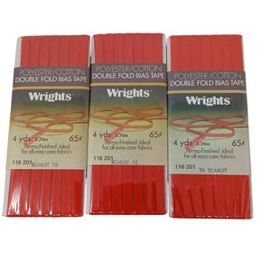 Op de afbeelding: Drie verpakkingen Wrights dubbele vouw biasband in scharlakenrood. Elke verpakking bevat 3,70 m polyester/katoenen band. De verpakking bevat de tekst "Perma-Finished - Ideal for all easy-care fabrics."