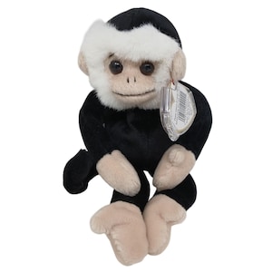 Peut inclure: Une peluche singe avec une fourrure noire et des mains, des pieds et un visage beiges. Le singe a une bordure de fourrure blanche autour de son visage et une étiquette attachée à son oreille. Le jouet est conçu pour être assis.