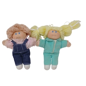May include: Two soft dolls with yarn hair and blue eyes. The left doll has peach-colored yarn hair, a pink checkered shirt, and dark blue denim overalls. The right doll has yellow yarn hair in pigtails, a light teal corduroy outfit, and white shoes.