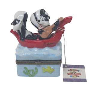Könnte beinhalten: Eine Looney Tunes Keramik-Schmuckschatulle mit Pepe Le Pew und einem weiteren Stinktier in einem roten Boot. Die Schatulle ist hellblau mit Wassermotiven und einer goldfarbenen Umrandung. Ein Looney Tunes-Etikett hängt an der Schatulle.