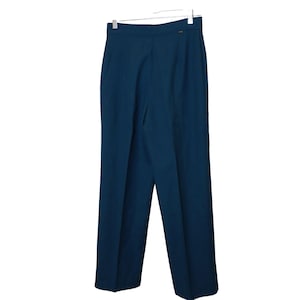 Puede incluir: Pantalones de color turquesa de cintura alta colgados de una percha blanca. Los pantalones tienen un diseño de pierna recta y un pequeño detalle dorado cerca de la cintura. La tela parece ser de color liso.