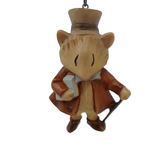 Może przedstawiać: Brązowa i beżowa figurka myszy w brązowym płaszczu, białej koszuli i cylindrze. Mysz trzyma mały biały pakunek i czarną laskę.