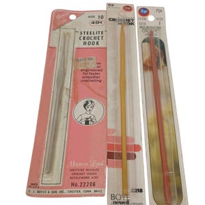 May include: Three vintage crochet hooks in their original packaging. The hooks are in clear plastic packaging with pink and yellow accents. One hook is silver, one is yellow, and one is orange.
