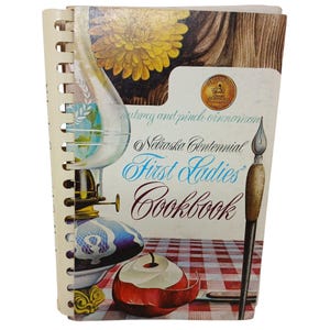 Może przedstawiać: Spiralnie oprawiona książka kucharska zatytułowana "Nebraska Centennial First Ladies Cookbook". Okładka zawiera ilustrację w stylu vintage przedstawiającą lampę, jabłko i pędzel. Tło ma wzór w kratę.