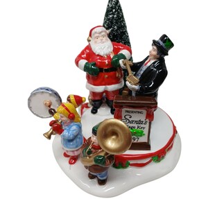 Può includere: Statuina natalizia con Babbo Natale, un uomo in smoking e bambini che suonano strumenti musicali. La base bianca presenta dettagli con nastri rossi e un piccolo albero di Natale verde.