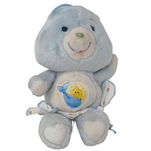 Puede incluir: Un peluche Care Bear azul claro con una barriga blanca con una estrella amarilla en una manta azul. El oso tiene una cara sonriente, detalles blancos y un lazo azul. Un juguete coleccionable vintage.