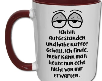 May include: White ceramic coffee mug with a maroon rim and handle. The mug features a cartoon illustration of two tired eyes and the text "Ich bin aufgestanden und habe Kaffee Geholt. Ich finde, mehr kann man heute nun echt nicht von mir erwarten."