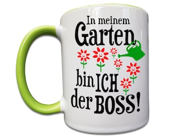Può includere: Tazza in ceramica bianca con manico verde. La tazza ha una grafica nera che dice "In meinem Garten bin ICH der BOSS!" con fiori rossi e un annaffiatoio verde.