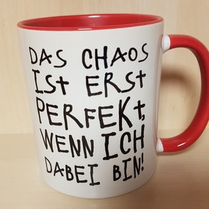 May include: White ceramic mug with a red rim and handle. The mug has black handwritten text that reads "Das Chaos ist erst perfekt, wenn ich dabei bin!"