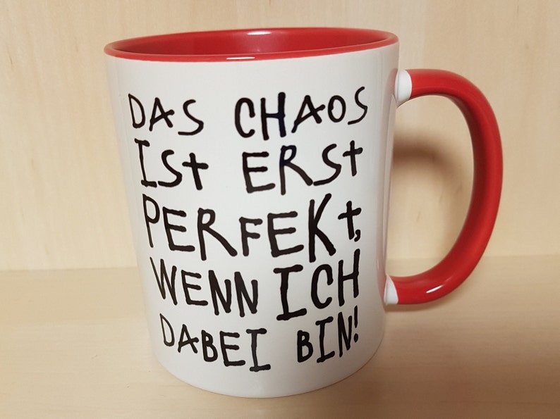 May include: White ceramic mug with a red rim and handle. The mug has black handwritten text that reads "Das Chaos ist erst perfekt, wenn ich dabei bin!"