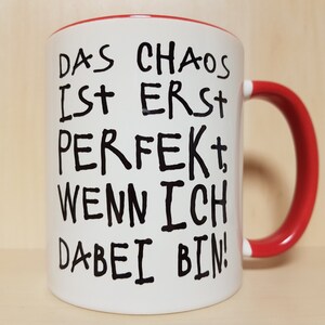 May include: White ceramic mug with a red rim and handle. The mug has black text that reads "Das Chaos ist erst perfekt, wenn ich dabei bin!"
