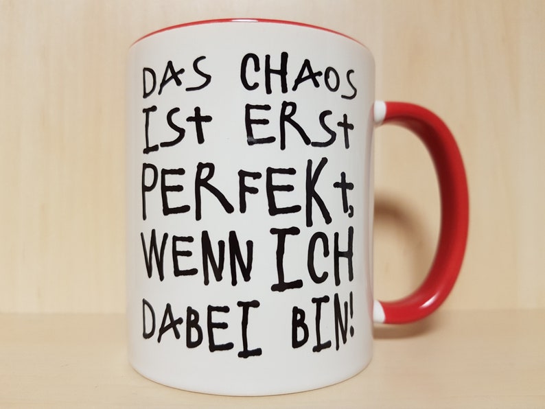 May include: White ceramic mug with a red rim and handle. The mug has black text that reads "Das Chaos ist erst perfekt, wenn ich dabei bin!"