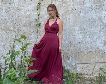 Może przedstawiać: Bordowa sukienka maxi z wiązaniem na szyi. Sukienka ma kopertowy gorset i zwiewną spódnicę. Modelka stoi przed teksturowaną betonową ścianą z zielenią. Sukienka jest w jednolitym kolorze i wydaje się być wykonana z miękkiej, zwiewnej tkaniny.