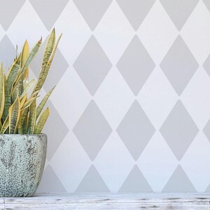 May include: A potted snake plant with long, green and yellow leaves sits in front of a wall with a repeating diamond pattern in light gray. The pot is a muted teal color and sits on a white wooden surface.