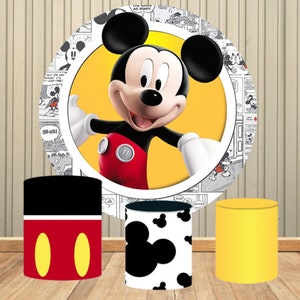 May include: Three round cardboard cake stands decorated with a Mickey Mouse theme. The largest stand features a cartoon image of Mickey Mouse in a red shirt and yellow shorts. The middle stand has a black and white pattern of Mickey Mouse's head. The smallest stand is black with red and yellow Mickey Mouse ears.