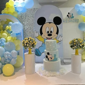 May include: A three-tiered cake with light blue and white patterns, featuring a Mickey Mouse graphic, sits on a white base. It is flanked by two yellow floral arrangements on white pedestals, with a large Mickey Mouse backdrop and blue and yellow balloons.