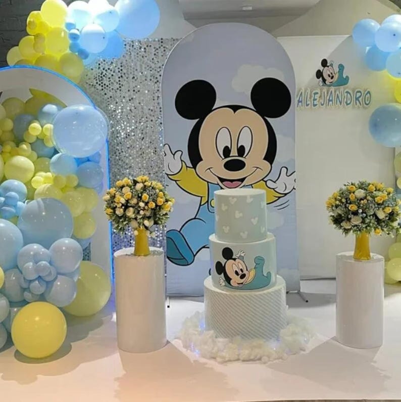 May include: A three-tiered cake with light blue and white patterns, featuring a Mickey Mouse graphic, sits on a white base. It is flanked by two yellow floral arrangements on white pedestals, with a large Mickey Mouse backdrop and blue and yellow balloons.