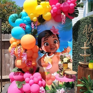 May include: A Moana themed party backdrop featuring a cartoon image of baby Moana. The backdrop is decorated with a garland of pink, orange, and blue balloons, green leaves, and pink flowers.
