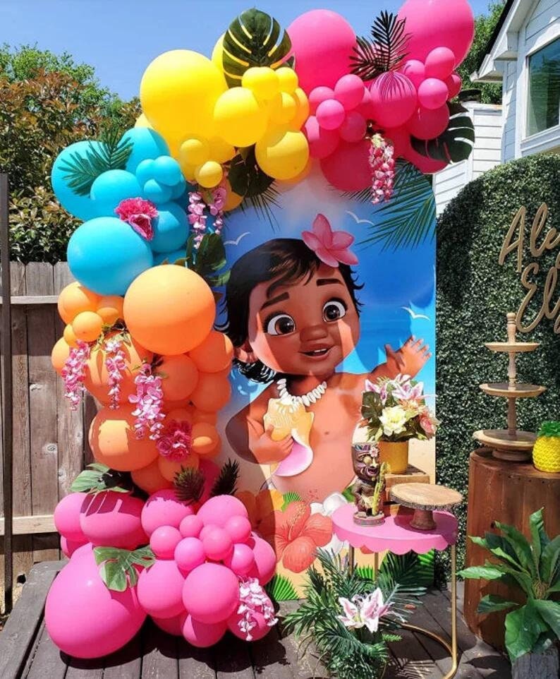 May include: A Moana themed party backdrop featuring a cartoon image of baby Moana. The backdrop is decorated with a garland of pink, orange, and blue balloons, green leaves, and pink flowers.