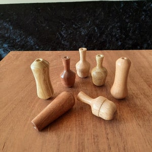 May include: A selection of seven wooden bottle stoppers in diverse forms and wood grains. The stoppers are arranged on a wooden surface, highlighting their unique designs and natural hues. The collection includes cylindrical, bulbous, and vase-shaped stoppers.