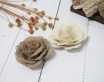 Könnte beinhalten: Zwei Blumen aus Burlap-Stoff, eine in hellbeige und eine in braun. Die Blumen liegen auf einer weißen Holzoberfläche.