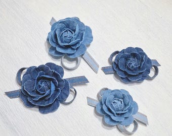 以下が含まれることがあります： デニム生地のフラワー装飾が4つ。各花は濃い青色で、バラに似ています。薄い青色のリボンとループに取り付けられています。花は白い表面に配置されています。