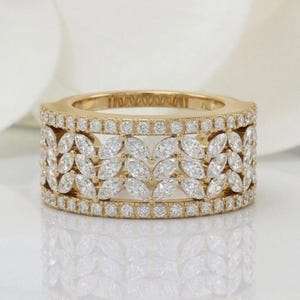 May include: A wide gold band ring featuring a central row of marquise-cut clear stones arranged in a leaf-like pattern, bordered by two thin bands of small, round, pavé-set clear stones along the top and bottom edges of the ring.